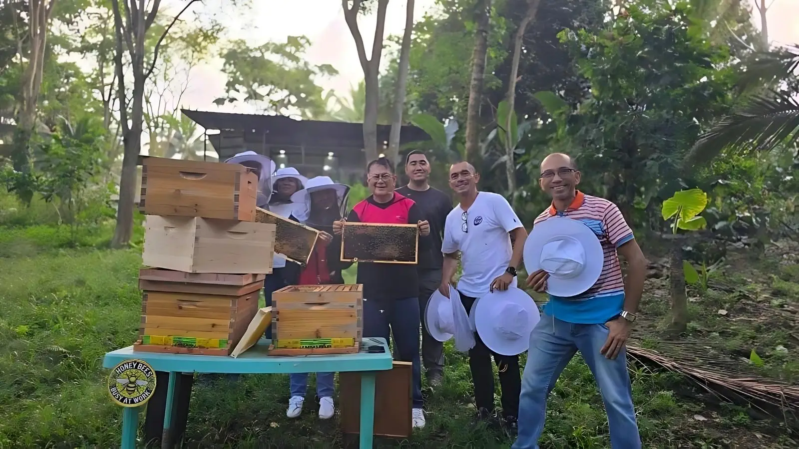 Beekeepers in protective gear inspecting honeycomb frames near wooden hives in a lush outdoor setting, demonstrating eco-friendly beekeeping practices at Honea Bee Farm.
