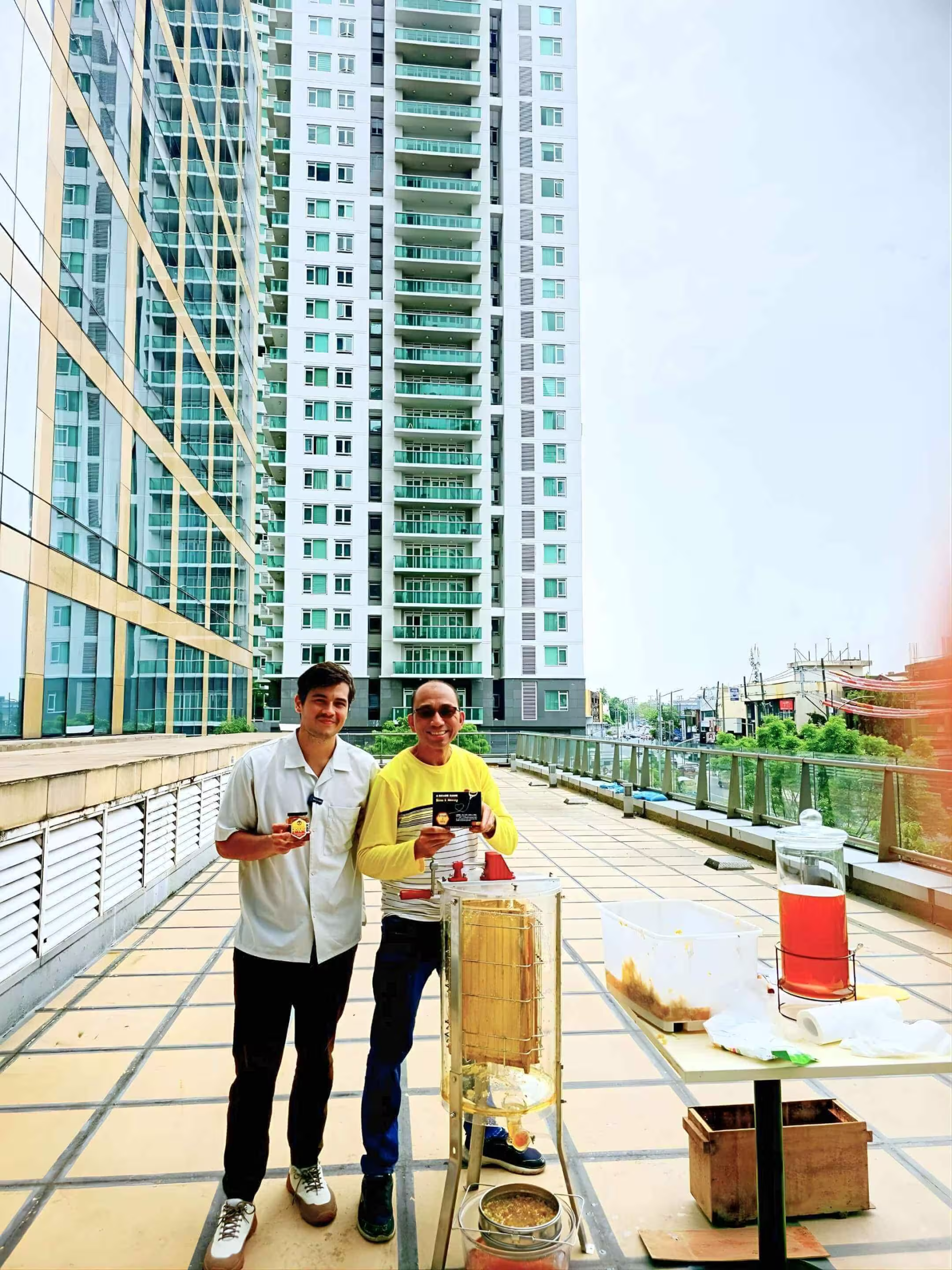 Two urban beekeepers on a city rooftop, holding jars of honey and a product box, next to a honey extractor and other beekeeping equipment with a high-rise building in the background.
